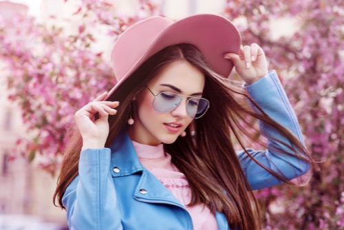 young-beautiful-fashionable-girl-wearing-stylish-blue-color-aviator-sunglasses--pink-suede-hat--earrings--biker-jacket.-model-posing-in-street-with-flowering-trees.-spring-fashion-concept