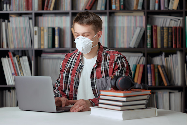 a-young-guy-in-a-surgical-mask-is-working-on-a-laptop-in-the-library-at-a-safe-distance-from-other-people